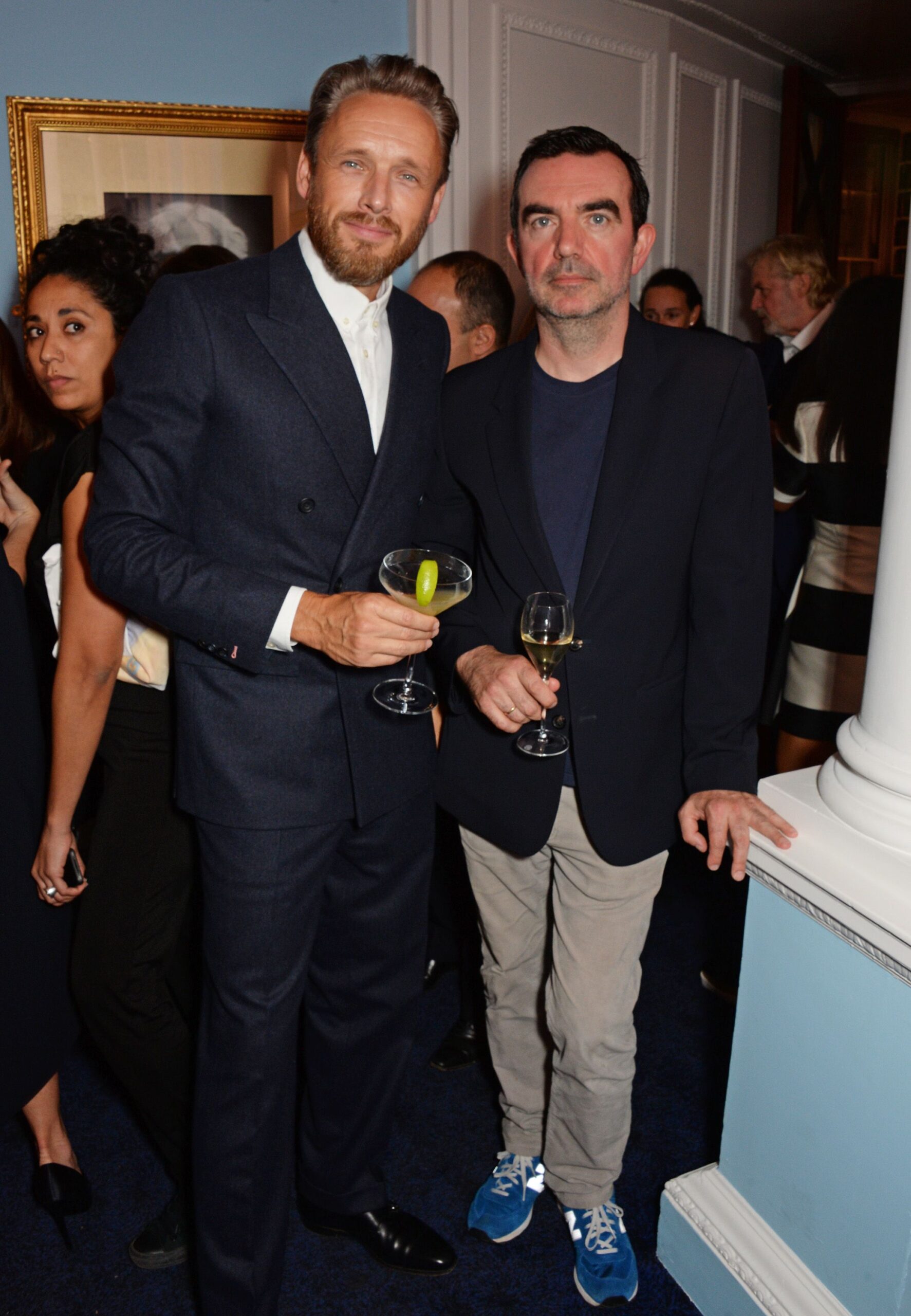 two men dressed in suits each holding a glass of wine, stood in front of a crowd of people