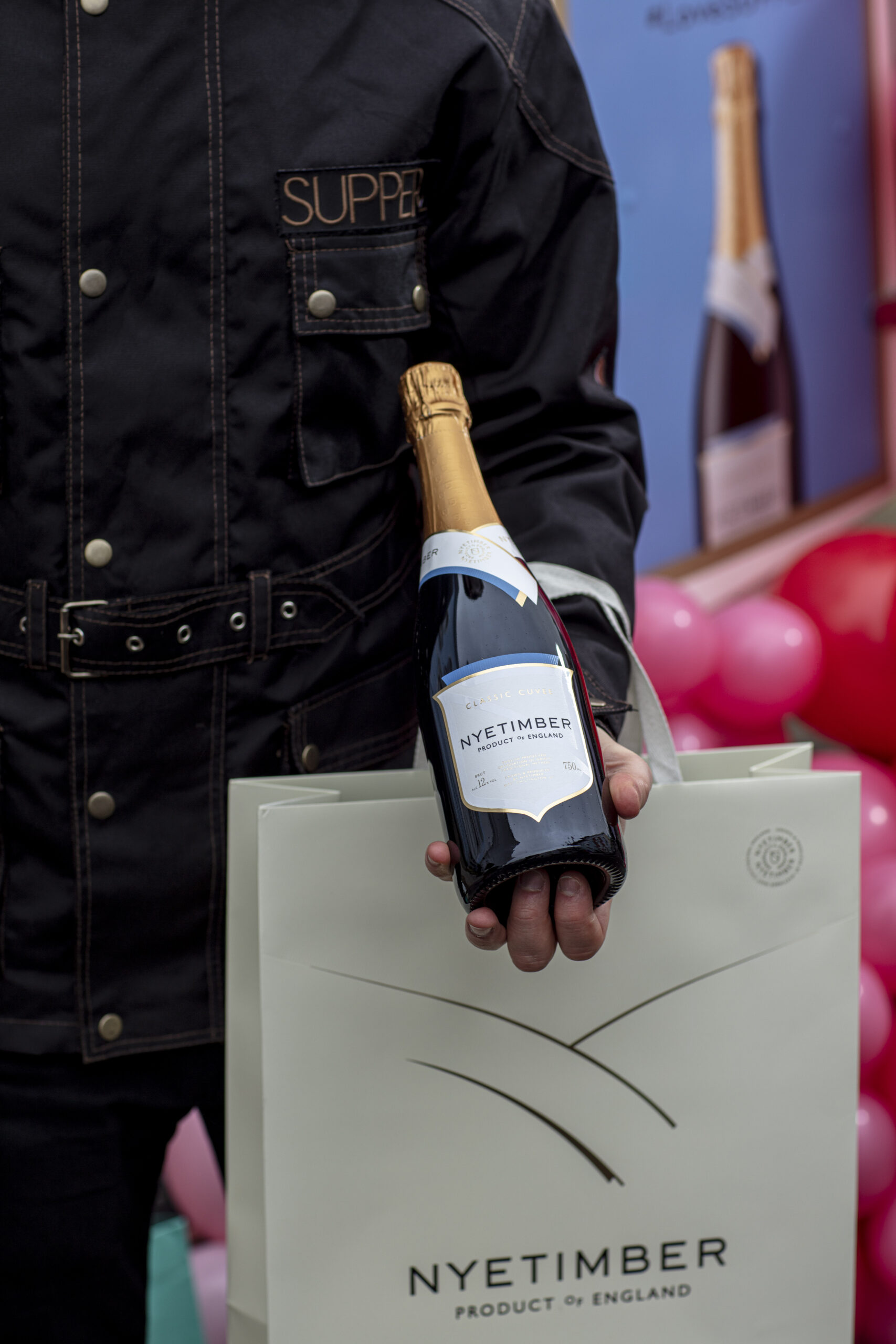 a man holding a bottle of Nyetimber wine in front of a branded Nyetimber bag