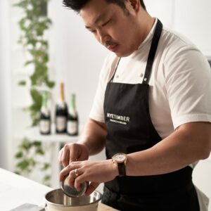 a chef pouring liquid into a pan with three wine bottles on a table in the background