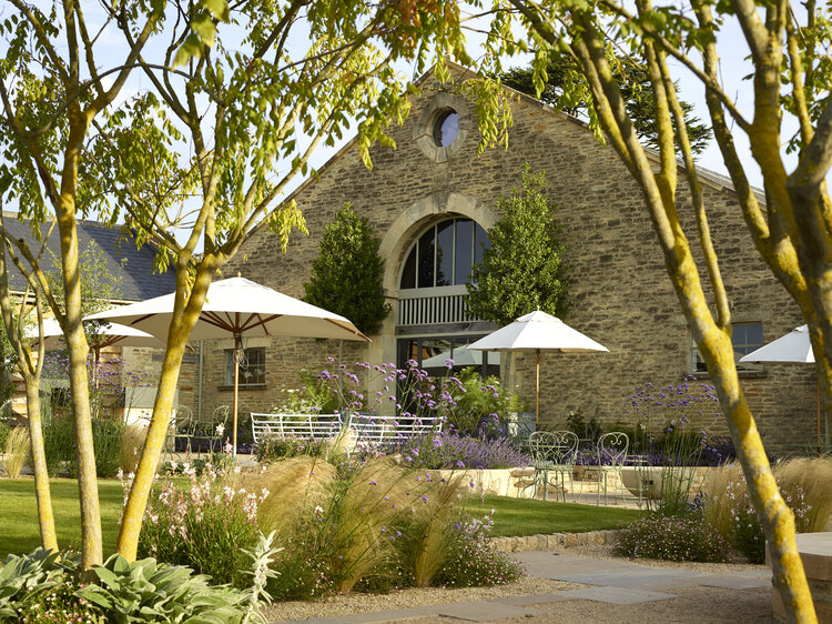 a garden with paving, lawn and tables set with white parasols in front of an exposed brick building with arched window